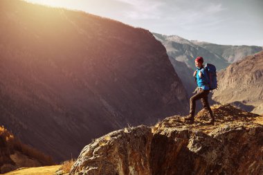 Sırt çantalı bir yürüyüşçü dağın tepesinde duruyor, elleri havada ve güneşin doğuşunun keyfini çıkarıyor.