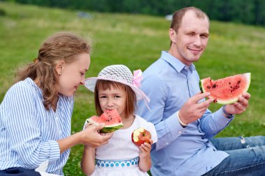 Piknikte karpuz yiyen mutlu bir aile. Anne, baba ve çocuk çayırda ya da parkta piknik yapıyorlar..