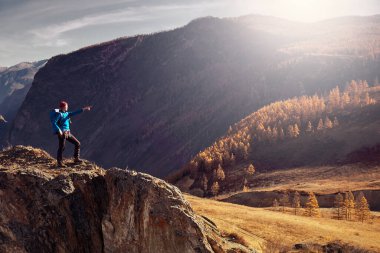 Sırt çantalı bir yürüyüşçü dağın tepesinde duruyor, elleri havada ve güneşin doğuşunun keyfini çıkarıyor.