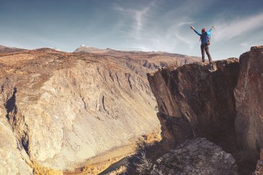 Sırt çantalı bir yürüyüşçü dağın tepesinde duruyor, elleri havada ve güneşin doğuşunun keyfini çıkarıyor.