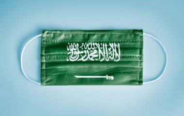 Covid-19 Coronavirus koruma konsepti: Mavi arka planda Suudi Arabistan bayrağı olan tıbbi tek kullanımlık yüz maskesi. WHO, Coronavirüs 'ü önlemek için sosyal uzaklık ve maske kullanımını tavsiye etti. 