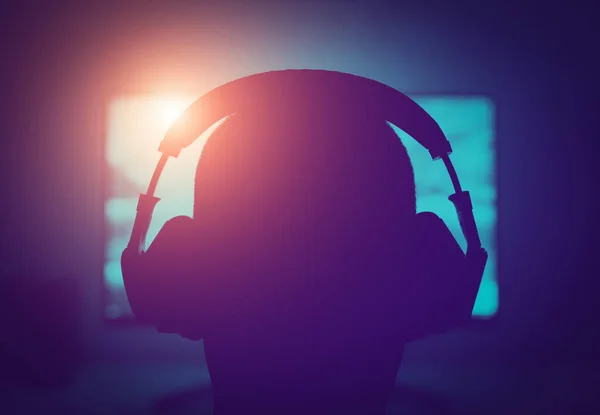 Young gamer with headphones playing computer game on glowing screen in a dark room. Video game addiction of young generation concept.