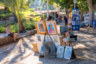 Rhodes Island-Yunanistan: 29 Ağustos 2024: Bir sanatçı güneşli bir parkta satılık diğer eserlerle çevrili bir palmiye ağacıyla bir portre çiziyor. Canlı atmosfer, popüler bir turistik bölgede yaratıcı bir ifade anı yakalıyor..