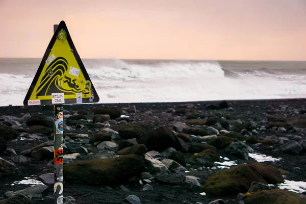 Um sinal que informa sobre os perigos na praia de Reynisfjara, com ondas esmagadoras no fundo — Fotografia de Stock