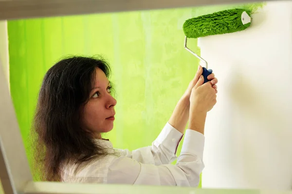 Pretty young woman in a white shirt is carefully painting green interior wall with roller in a new home, view from behind through a ladder