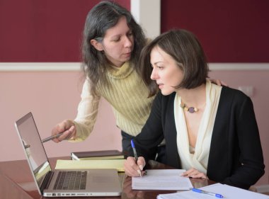 Zeki giyimli genç bir bayan başka bir genç bayanla sorunları tartışıyor, dizüstü bilgisayarı işaret ediyor ve bir çözüm öneriyor, klasik ofis odası, klasik ofis masası.