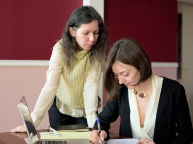 Zeki giyimli genç bir bayan başka bir genç bayanla sorunları tartışıyor, dizüstü bilgisayarı işaret ediyor ve bir çözüm öneriyor, klasik ofis odası, klasik ofis masası.