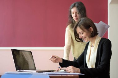 Genç ve şık giyimli bir bayan başka bir genç bayana evraklarla çalışması için yardım ediyor, formları dolduruyor ve imzalıyor. Eski tarz bir ofiste, eski bir masada birlikte oturuyorlar.