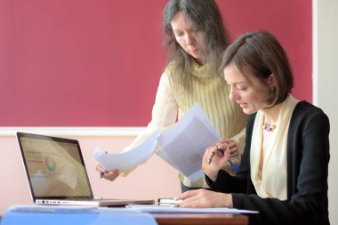 Genç ve şık giyimli bir bayan başka bir genç bayana evraklarla çalışması için yardım ediyor, formları dolduruyor ve imzalıyor. Eski tarz bir ofiste, eski bir masada birlikte oturuyorlar.
