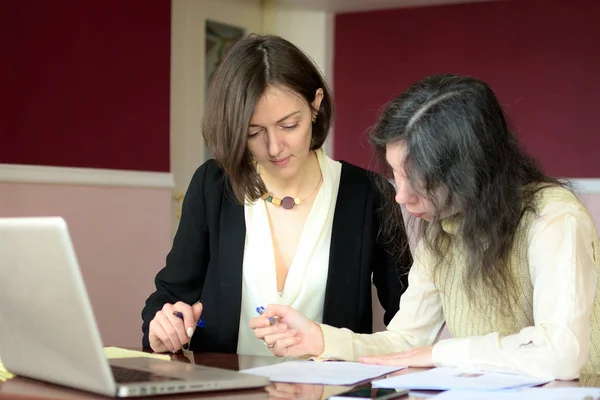 Joven dama elegantemente vestida ayuda a otra joven a trabajar con documentos, llenar formularios y firmar. Se sientan juntos en una oficina de estilo vintage en un escritorio vintage Imagen De Stock