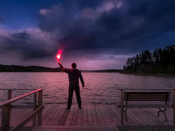 A man at the lake lit the signal block. SOS. Help signal. The man at the pier is holding signal block. The man on the quay. Torch with fire.