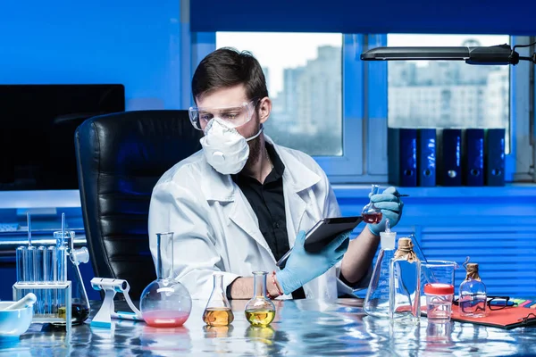 Man works as a laboratory assistant. Laboratory assistant with test tubes in hands. Human works in a chemistry lab. Man in a protective mask works with chemicals. Lab assistant with a tablet in hands