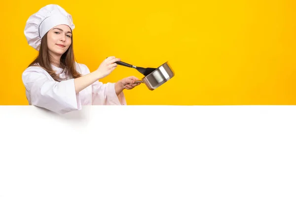 Shef girl on a yellow background. Female cook smiles and stirs something. Woman in a cook suit. Restaurant chef next to a white banner. Concept - chef recommended. Work as a cook in a restaurant