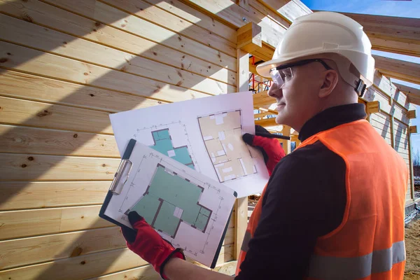 Building bussiness. Builder holds drawings of house in his hands. Human is considering a house plan on paper. Man in a protective helmet. Builder next to the frame of building. Wooden house frame