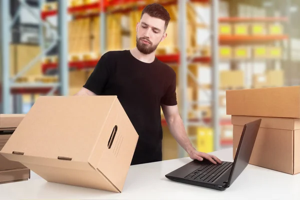 A bearded man against the background of warehouse shelves. Storekeeper with a laptop and cardboard boxes. Working in a warehouse. Storage of goods.