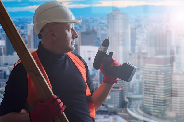 Repair of apartments. Home improvement. A man in a construction helmet on the background of the city. A man with a Board and an electric screwdriver in profile against the background of houses.