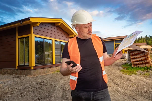 Man near a newly built cottage. Man is dissatisfied with the built house. Dissatisfaction with the work of builders. Builder examines drawings. Concept - work as a team leader at a construction site