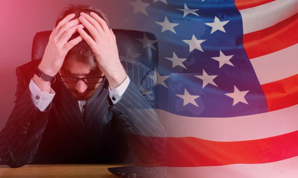 Upset man next to USA flag. Guy is in depression. Man is sitting and holding on to his head. Concept - man is saddened by situation in America. Disorder happening in USA. Concept - bad news from USA.