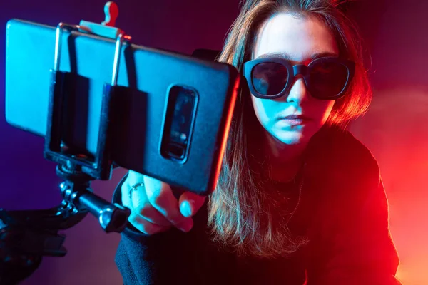 Girl video blogger. Phone mounted on a tripod in front of a woman. Girl student in dark. Young woman takes pictures of herself on phone. Video blogger is recording broadcast. Neon lighting.