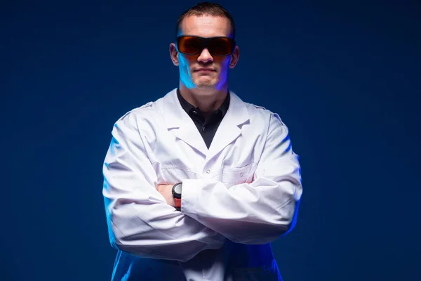 Doctor hero. A man in a medical gown stands with his arms crossed. Medic poses against a dark background. Portrait of a doctor in sunglasses. A large man in a doctor\'s uniform. Medic looking camera