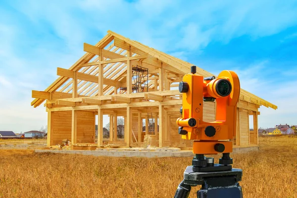 Optical level next to the wooden frame. Concept - frame quality check. Theodelite mounted on a tripod. Kotsnept - the use of theodelite in construction. Wooden house frame on blue sky background