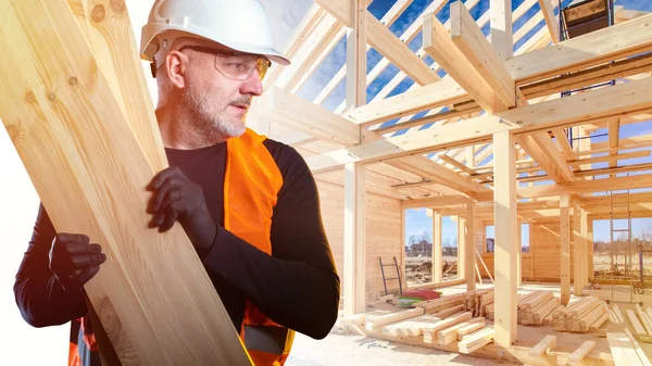 Builder carries several boards. Man works at a construction site. Man is  building a house. Builder near frame of the house. Construction of the  house. Concept - sale of frame houses. Career
