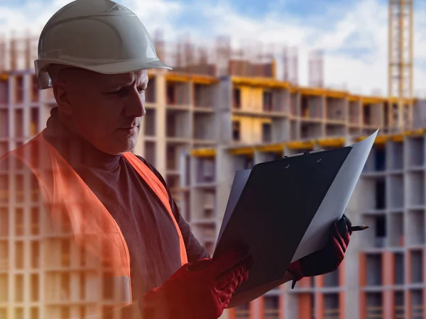 Builder holds a clipboard in hands. Builder on background of high-rise buildings under construction. Construction of high-rise buildings. Concept - architect. A man is considering something.