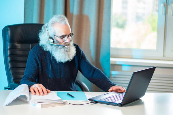 Experienced employee. A man with gray hair and a beard is working at a computer. An elderly man in headphones with a microphone in the office.