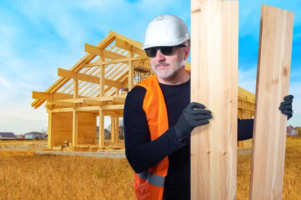Building wood house. Builder next to frame of wooden cottage. Skeleton of wooden house on sky background. Man carries timber for construction. Use of wood for building. Builder in hard hat with beams