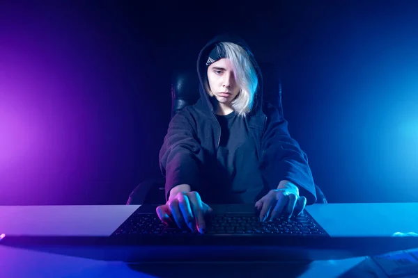 Woman hacker in a neon light. Cybercriminal. Hacker holds hands on keyboard. Hacking computer networks. Concept - committing cyber crime. Girl in hood looks at laptop. Hacker front view. Hacking.