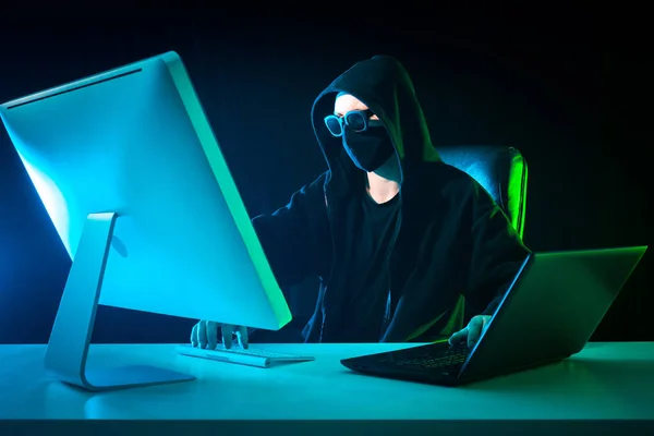 Hacker near computer. Anonymous. Man hiding his face works a computer. Paren breaks into computer networks. Hacker uses two laptop a time. Computer cracker in a dark mask. Anonymous hack security.