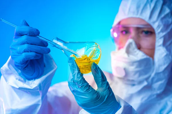 A flask with a yellow chemical liquid in the hands of a chemist. Laboratory assistant in protective clothing holding a flask. Virologist. Ecologist. The girl mixes the chemical liquid.