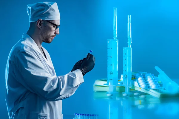 Doctor drug testing. Pills and syringes next to doctor. Medic on a blue background. Concept - drug testing in laboratory. Loborant examines test tube with a vaccine. Vaccine testing. Drug production