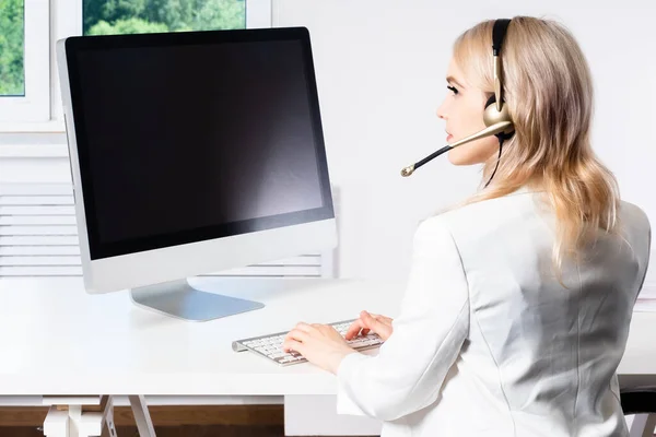 A girl in a white suit and headphones. A woman works at a computer. Telecommuting. The blonde consults clients through a computer application. A beautiful girl is sitting at her Desk.