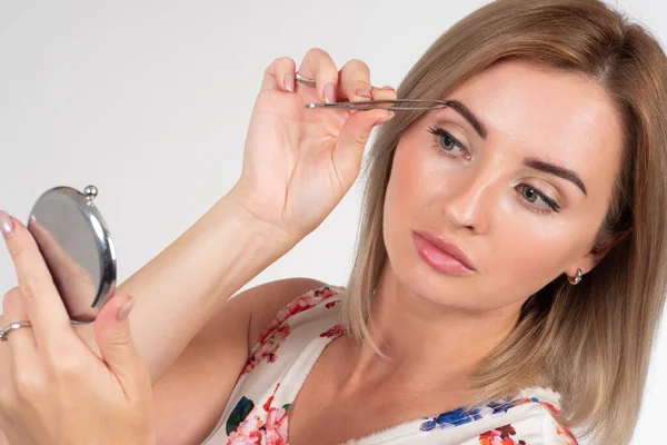 Woman plucking her eyebrows with tweezers. Girl does her makeup. Woman with tweezers on a light background. Woman with tweezers and a mirror in her hands. Eyebrows removal. Laser. Makeup.