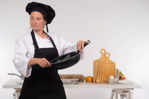 Female chef. Chef with a frying pan in his hands. A woman in a chef costume on the background of the kitchen. A woman with a frying pan in her hands looks away.