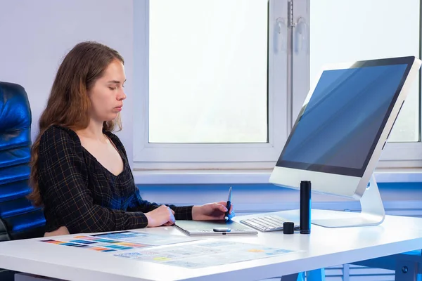 Graphic designer is working. Woman in office draws on graphics tablet. Girl next to computer. Concept - she pursuing a career as a graphic designer. Woman is developing design of mobile applications.