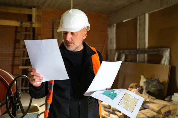 A man in a white helmet inside a building under construction. The Builder studies the drawings. Major repairs in the house. A man in construction clothes. Repair of apartments and offices.