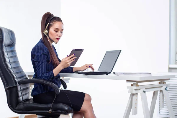 Hard work. A girl in a business suit at a Desk. A woman in headphones with a microphone. The girl uses a computer and tablet in her work. Focused worker.