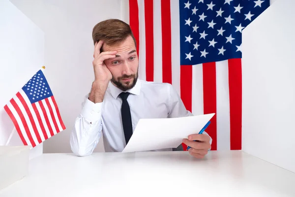 American official reads the documents. A man with documents on the background of the US flag. A person is amazed when reading American documents. A man fills out papers for immigration to the USA.