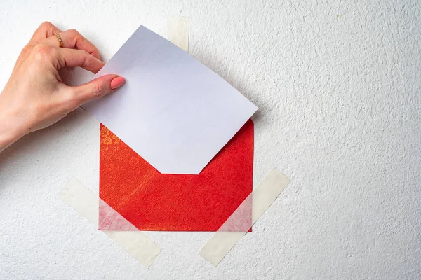 Design background with a red envelope and a piece of paper. The girl puts a piece of paper in an envelope. The red envelope is taped to the wall. Letter. Message. Note.