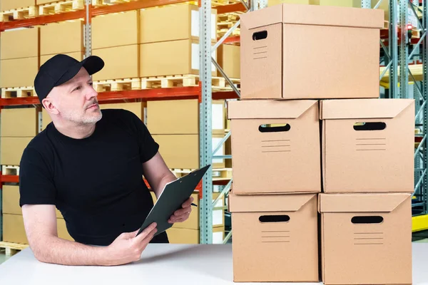 Warehouse worker on background of shelves. Warehouse company employee next to boxes. Man is holding a clipboard. Concept - a career of a warehouse worker. Corton boxes next to storehouse worker.