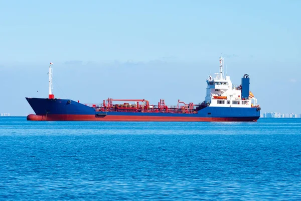 Tanker on the ocean backdrop. Chemical tanker is sailing on sea. Ship for transportation of chemical products. City can be seen in the distance. Chemical tanker is painted blue and red.