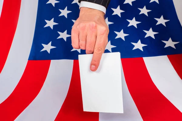 Man hand with a white ballot paper. Voter hand on background of flag of America. Concept - a place for inscription on ballot paper. Blank ballot in American hand. Voting in USA. Man votes. Referendum