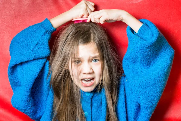 Mignonne fille dans un pull bleu se brosse les cheveux sur un fond rouge — Photo