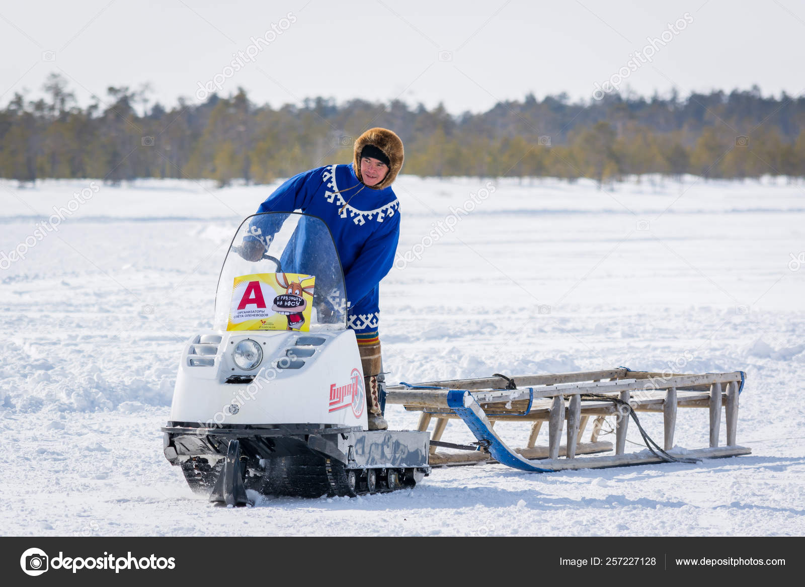 Russkinskaya Russia 24 March 18 Taiga Resident Of Siberia In National Northern Clothing Riding Snowmobile With Wooden Sledges In The Snow Against The Background Of Winter Forest Holiday Of Th