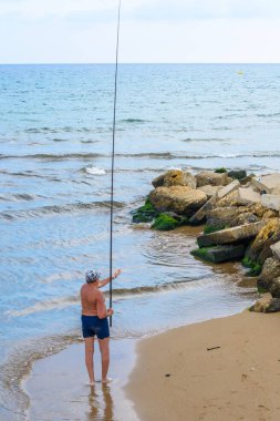 Side, Türkiye - Haziran 2018: Akdeniz sahilinde uzun olta ile mayo ve şapkalı tabaklanmış adam.