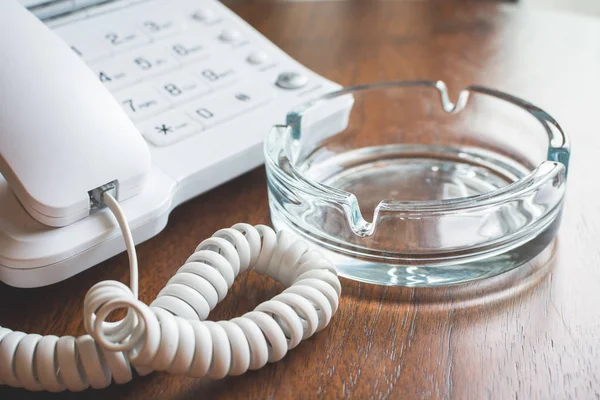 Telefone branco nos botões ao lado de um cinzeiro de vidro em uma mesa de madeira . — Fotografia de Stock