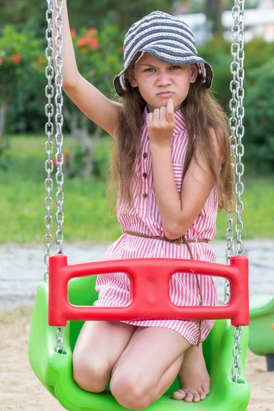 Side, Turquia - Junho de 2018: Menina bonita em um chapéu e um vestido listrado com um olhar inteligente está sentado no balanço do parque infantil . — Fotografia de Stock