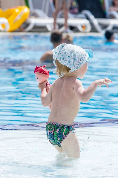 Little child in shorts and cap scares people holding the doll in the hand of the blue pool.